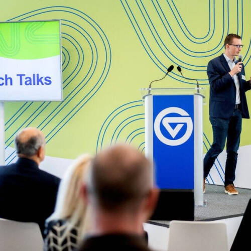 Person in a dark blazer and jeans speaks into a microphone at a podium branded with the “GVSU” logo during a “GVSU Tech Talks” event, while an audience listens from modern white chairs. A large screen beside the speaker displays “GVSU Tech Ta...